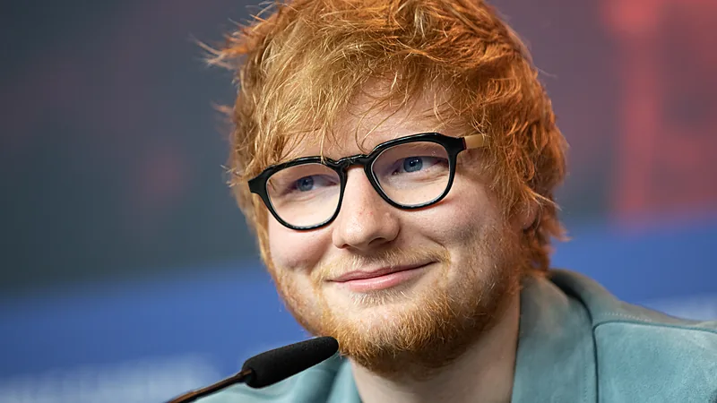 Ed Sheeran tersenyum berkacamata di depan mikrofon pada sebuah konferensi pers