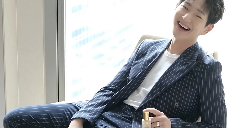 ONEW tertawa mengenakan setelan bergaris duduk bersandar di kursi dekat jendela