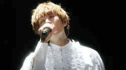 ONEW SHINee bernyanyi memegang mikrofon dengan kemeja putih berenda di atas panggung konser