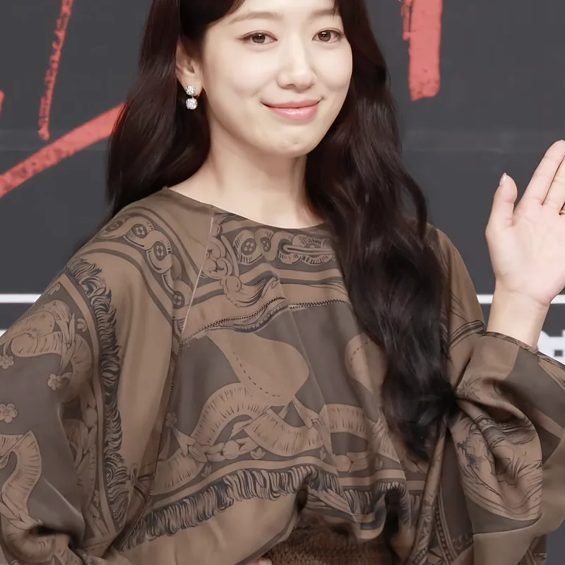 Park Shin-hye tersenyum melambaikan tangan dengan blus cokelat bermotif