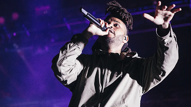 The Weeknd bernyanyi memegang mikrofon dengan jaket abu-abu di atas panggung