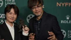 Ryunosuke Kamiki memegang figur Godzilla berdiri bersama Takashi Yamazaki di acara karpet merah, dengan teks Godzilla Minus Zero