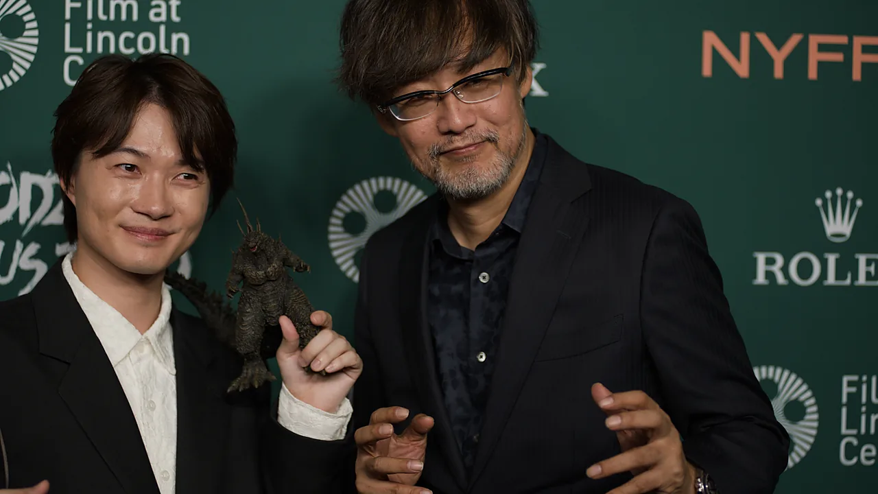 Ryunosuke Kamiki memegang figur Godzilla berdiri bersama Takashi Yamazaki di acara karpet merah, dengan teks Godzilla Minus Zero