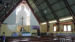 Bagian dalam gereja katolik dengan salib kayu besar di dinding altar dan deretan bangku jemaat