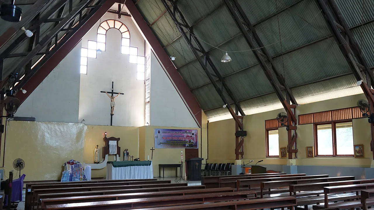 Bagian dalam gereja katolik dengan salib kayu besar di dinding altar dan deretan bangku jemaat